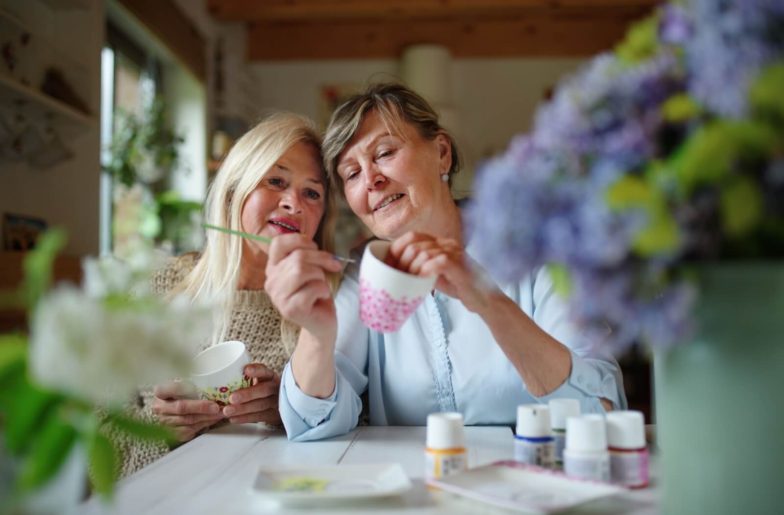 two mature friends in assisted living use paint to decorate ceramic cups.