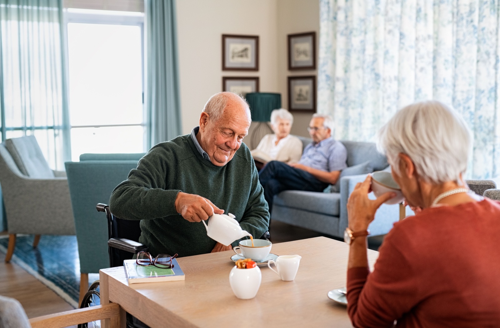 A senior couple in a memory care community sit at a table drinking tea.