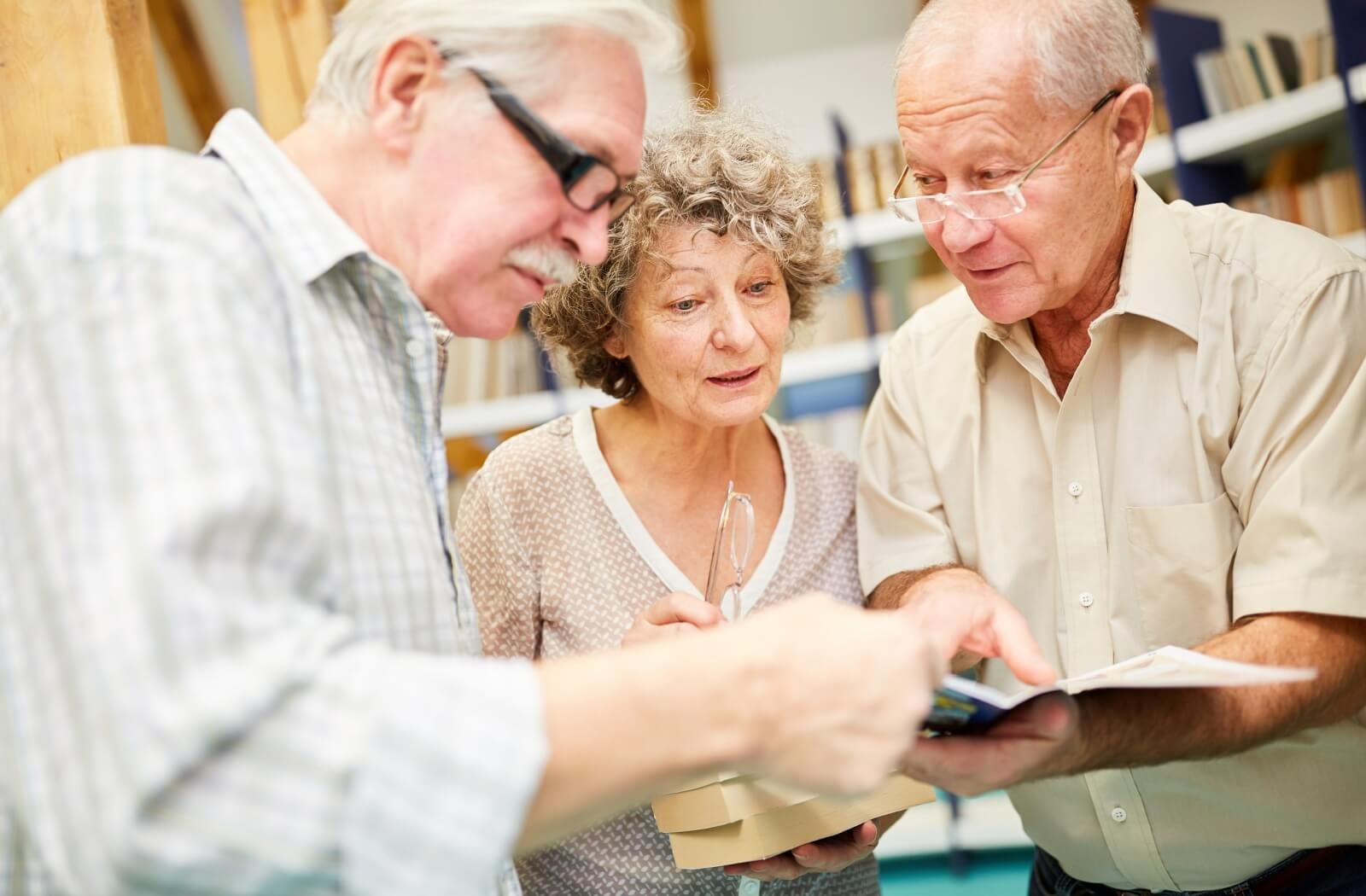 3 older friends examine books in a library, learning together and building social connections.
