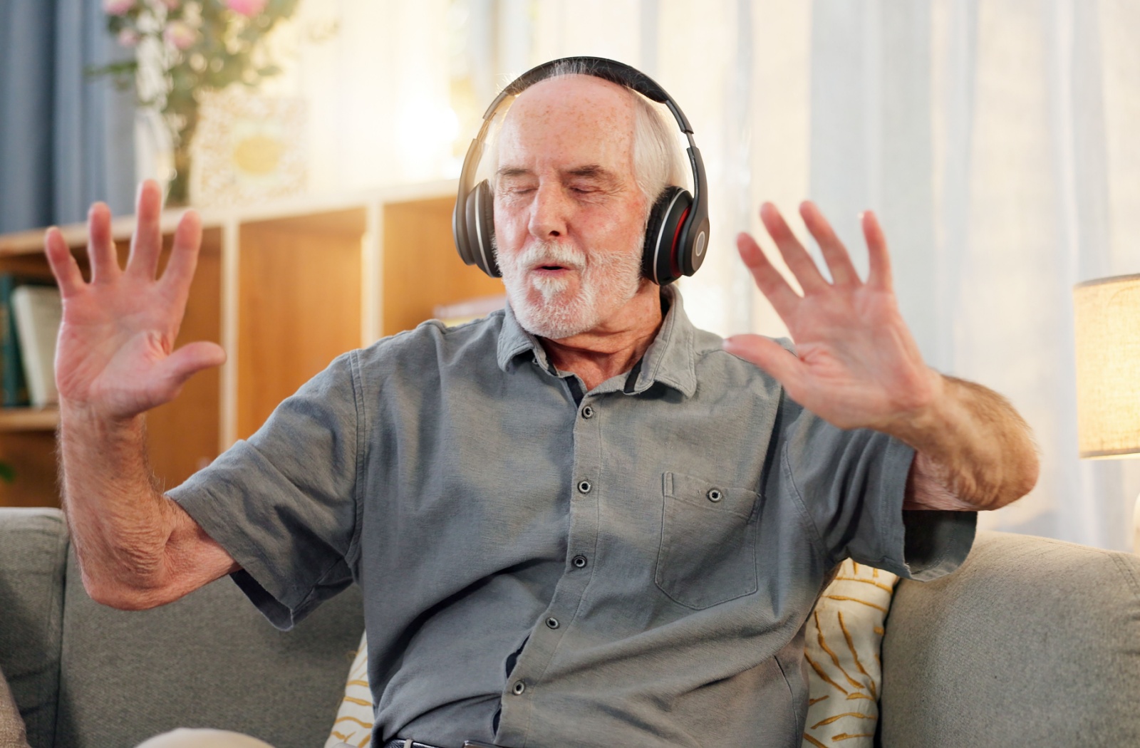 An older adult dances to music while sitting on their couch, wearing headphones in their home in senior living
