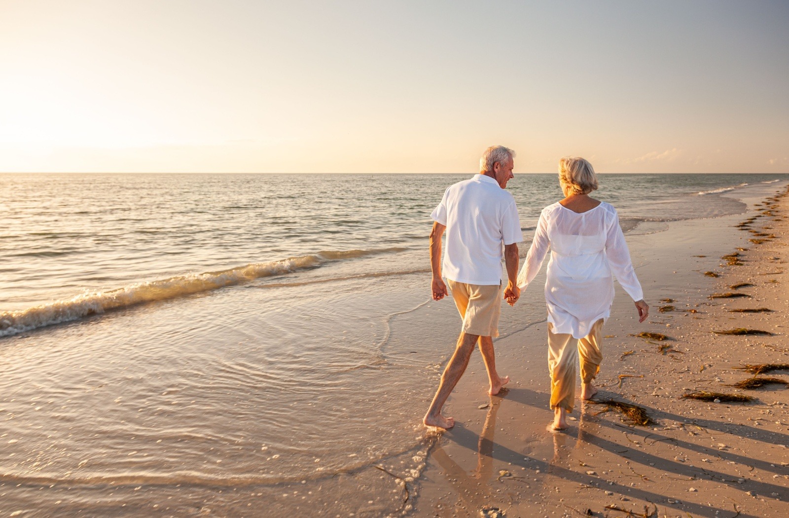 An older couple holds hands and enjoys a stroll along a beach during sunset
