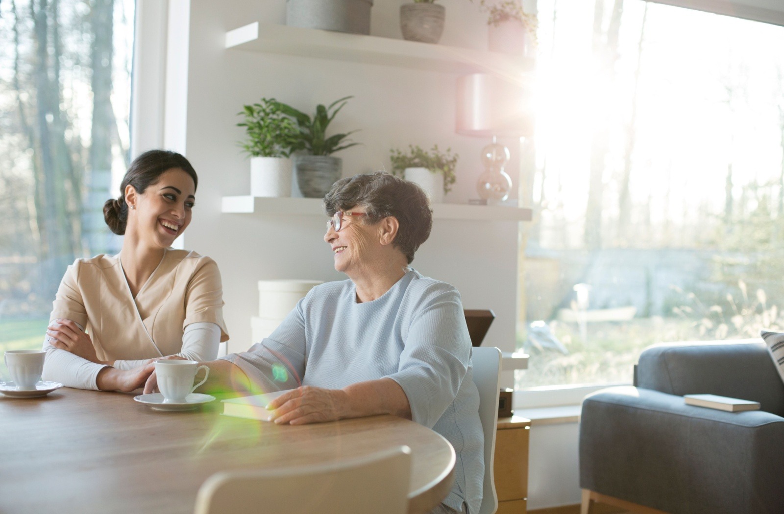 An adult child and their senior parent smile as they reminisce while seated at a kitchen table.
