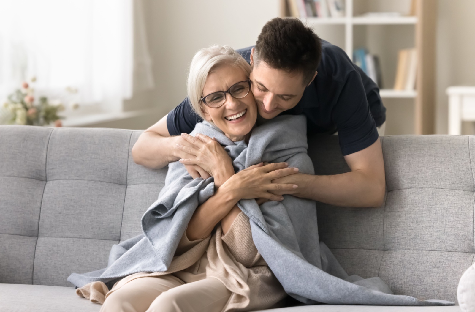 a senior resident sits on a gray couch while wrapped in a blanket and being embraced by their adult child.