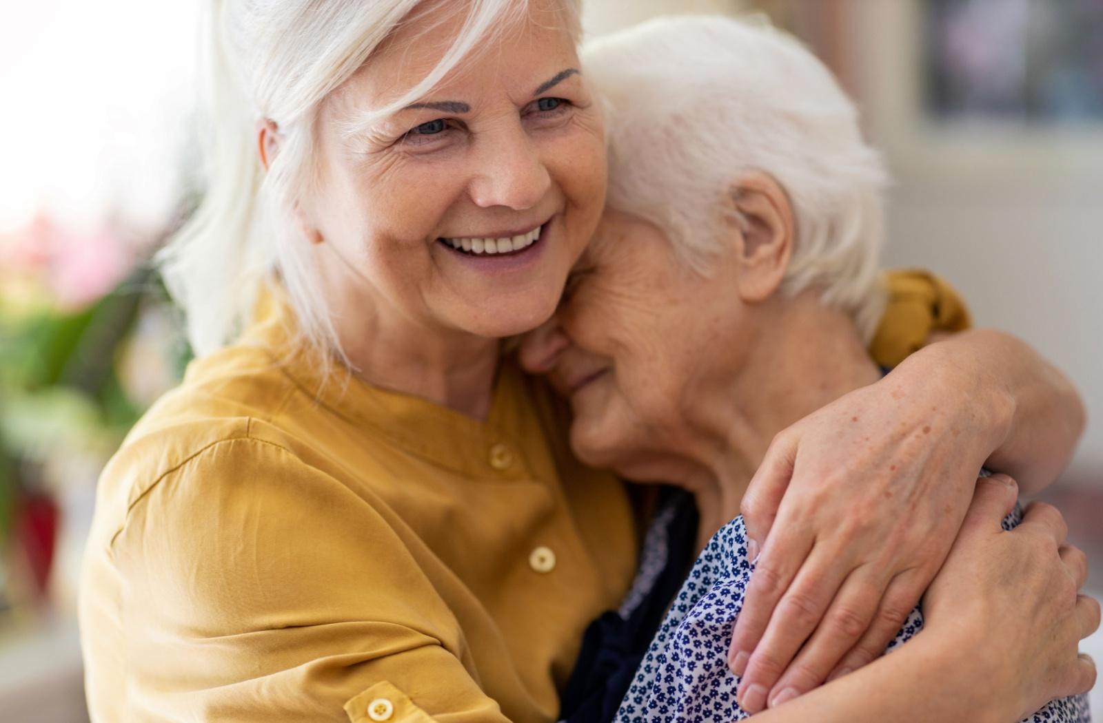A close-up of a middle-aged individual in a mustard-yellow shirt affectionately hugging a senior, who has their eyes closed in comfort.